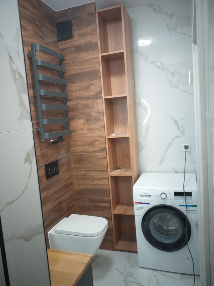 Nowoczesna łazienka z płytkami imitującymi drewno i marmur, grzejnik drabinkowy, podwieszana toaleta i pralka marki Bosch, wysoka drewniana szafka z półkami.
