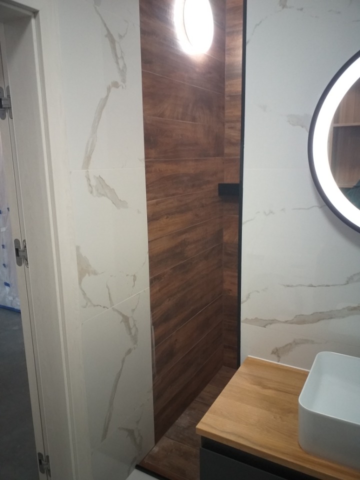 Nowoczesna łazienka z kabiną prysznicową wyłożoną płytkami imitującymi drewno, kontrastująca z białymi płytkami z marmurowym wzorem na pozostałych ścianach, okrągłe lustro z podświetleniem LED...