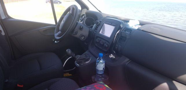 Wnętrze busa z widokiem na morze, butelka wody na konsoli środkowej, ciemna tapicerka.