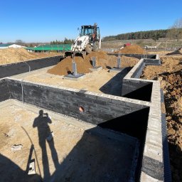 Usługi Koparko - Ładowarką Ponicki Radosław - Fundamenty z bloczków betonowych, częściowo zasypane ziemią, z koparką w tle. Widoczny cień osoby robiącej zdjęcie. Słoneczny dzień, teren budowy.