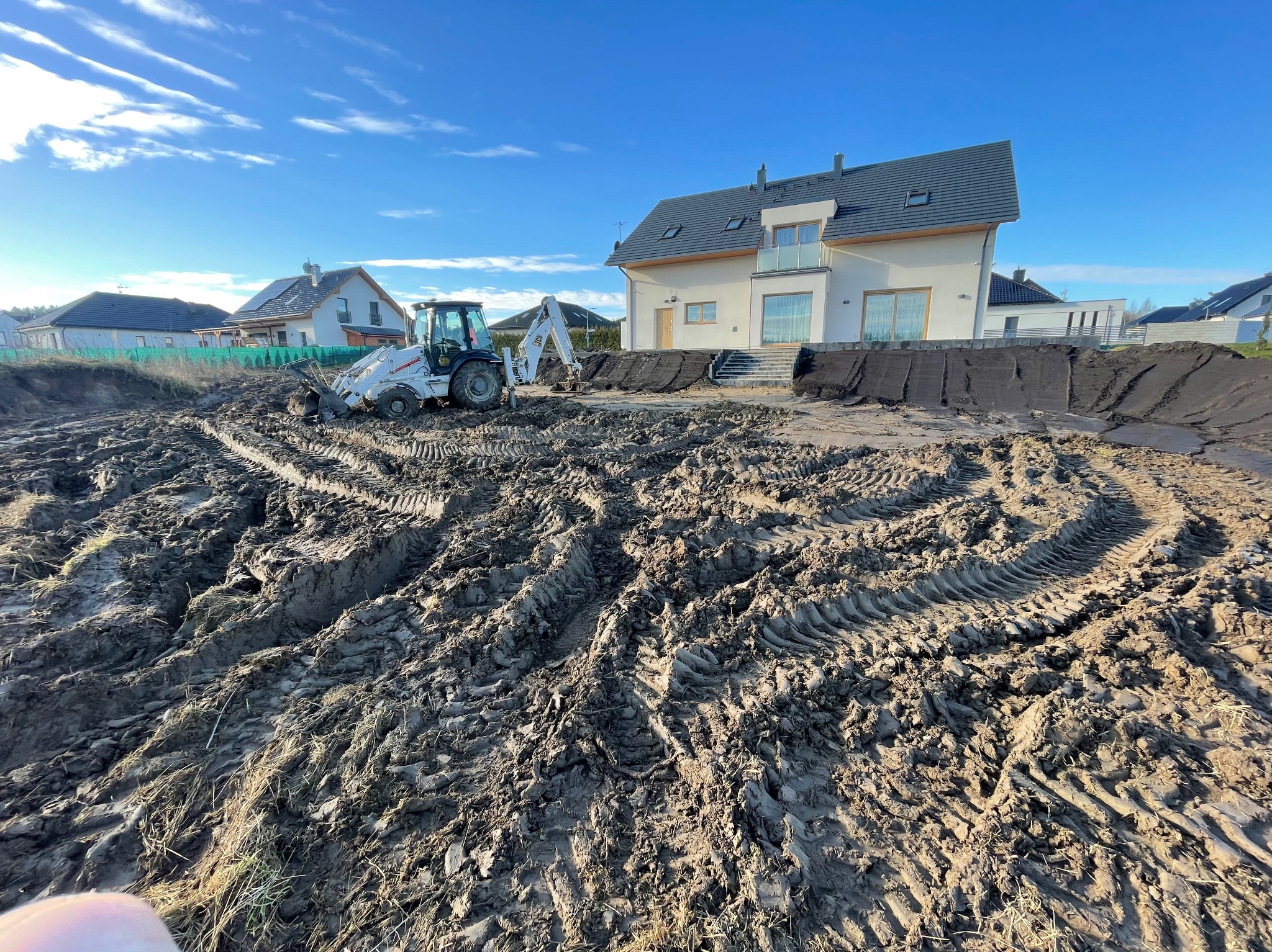 Teren przed niwelacją: koparko-ładowarka na nierównym, błotnistym placu budowy przed nowoczesnym domem z szarą dachówką i jasną elewacją, błękitne niebo.