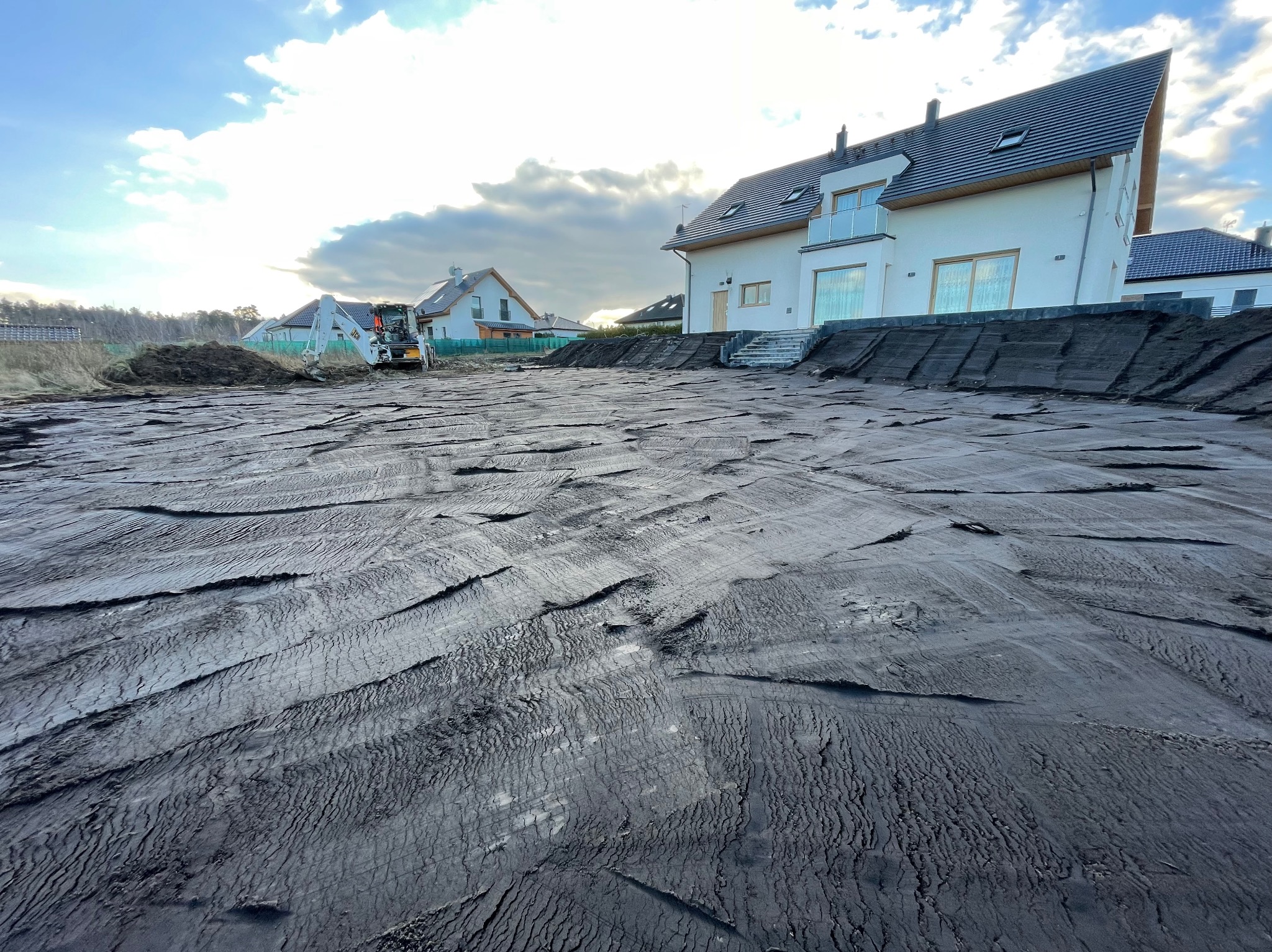 Nierówny teren po niwelacji, z widocznymi śladami opon i koparką w tle, przed nowym, białym domem z ciemnym dachem w słoneczny dzień.