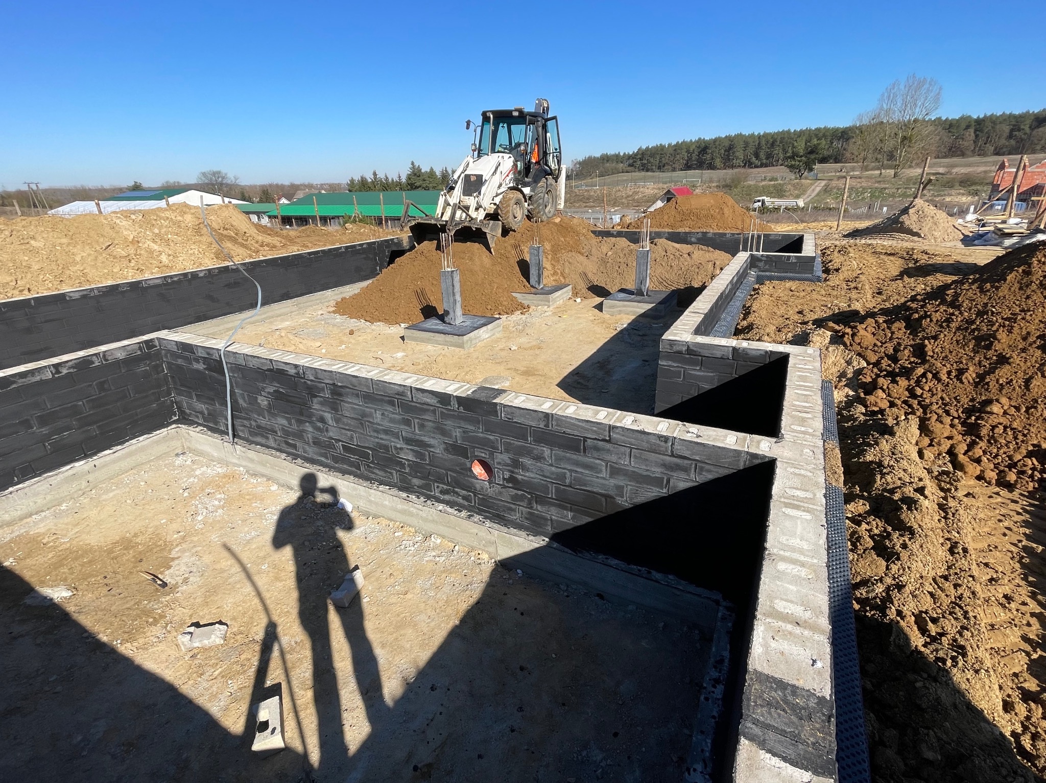 Fundamenty z bloczków betonowych, częściowo zasypane ziemią, z koparką w tle. Widoczny cień osoby robiącej zdjęcie. Słoneczny dzień, teren budowy.