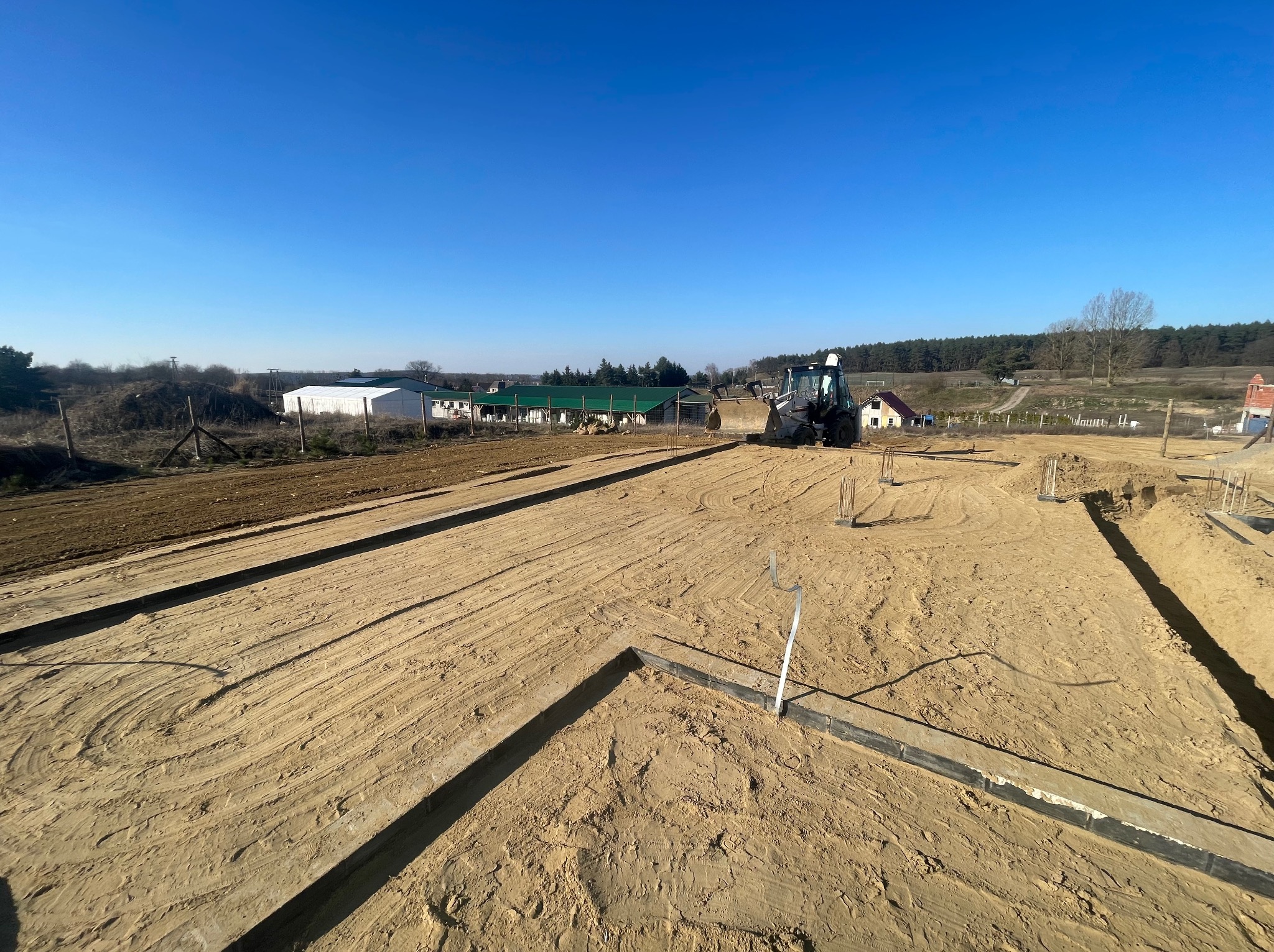 Przygotowany teren pod fundamenty z wykopanymi rowami, widoczny miniładowarka i częściowo zniwelowany teren. Słoneczny dzień, błękitne niebo.