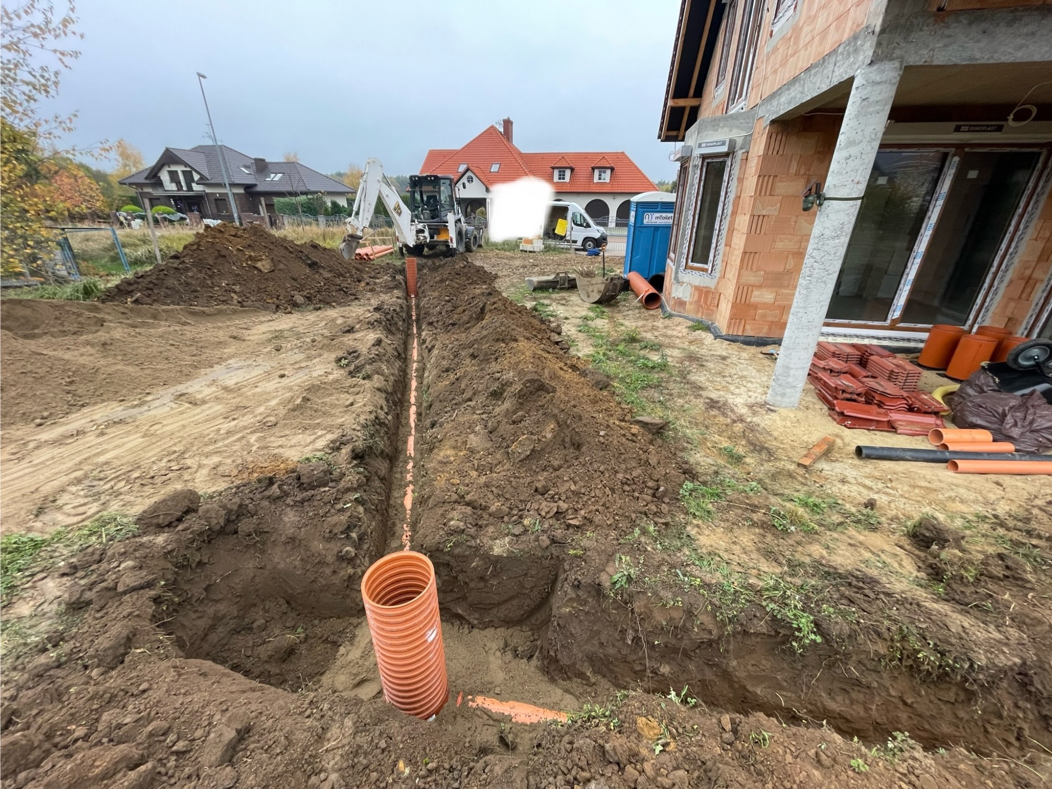 Wykop z ułożonymi rurami kanalizacyjnymi, wykonany koparką na tle budowy domu jednorodzinnego i przenośnej toalety. Widoczny stos dachówek.