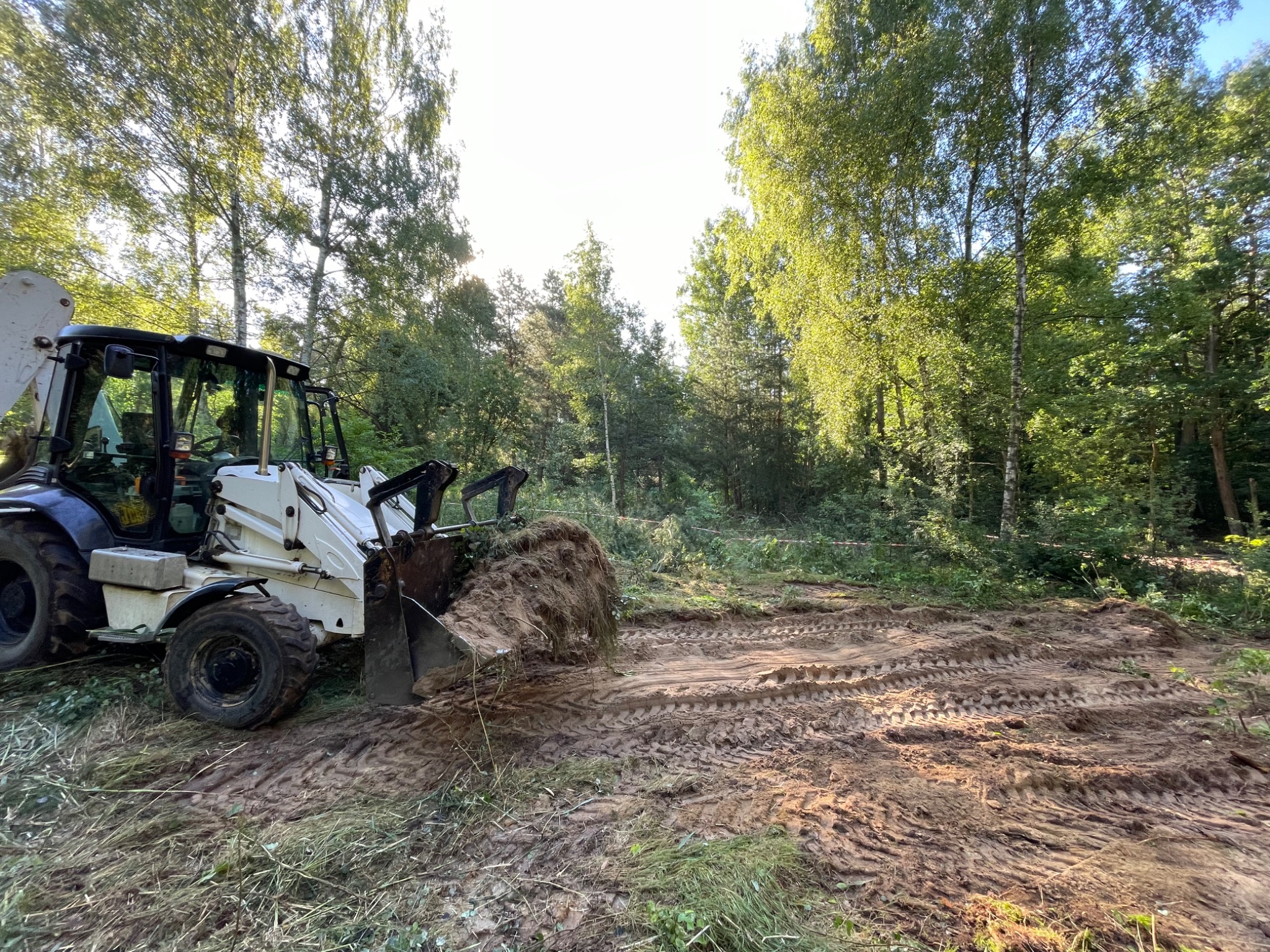 Biała koparko-ładowarka w trakcie niwelacji terenu leśnego. Ślady opon na ubitej ziemi, drzewa w tle. W tle zielone drzewa i krzewy.