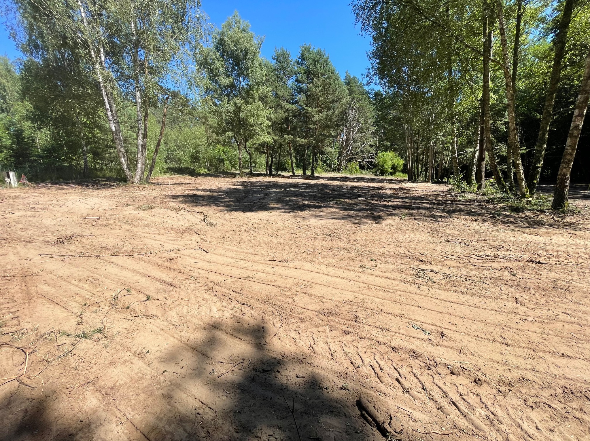 Wyrównany, piaszczysty teren otoczony drzewami w słoneczny dzień, z widocznymi śladami opon, sugerujący przygotowanie pod dalsze prace budowlane lub zagospodarowanie.