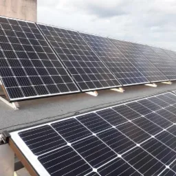 Rząd paneli słonecznych zamontowanych na płaskim dachu, widoczne elementy mocujące i częściowo widoczne tło z drzewami i budynkami.