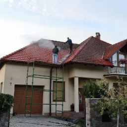 Dwóch mężczyzn na rusztowaniu i drabinie czyści dach z czerwonej dachówki za pomocą myjki ciśnieniowej, widoczny dom z garażem i balkonem z kwiatami.