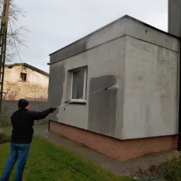Mężczyzna w czarnej kurtce czyści elewację budynku z szarego tynku za pomocą myjki ciśnieniowej, widoczny efekt różnicy czystości przed i po myciu.