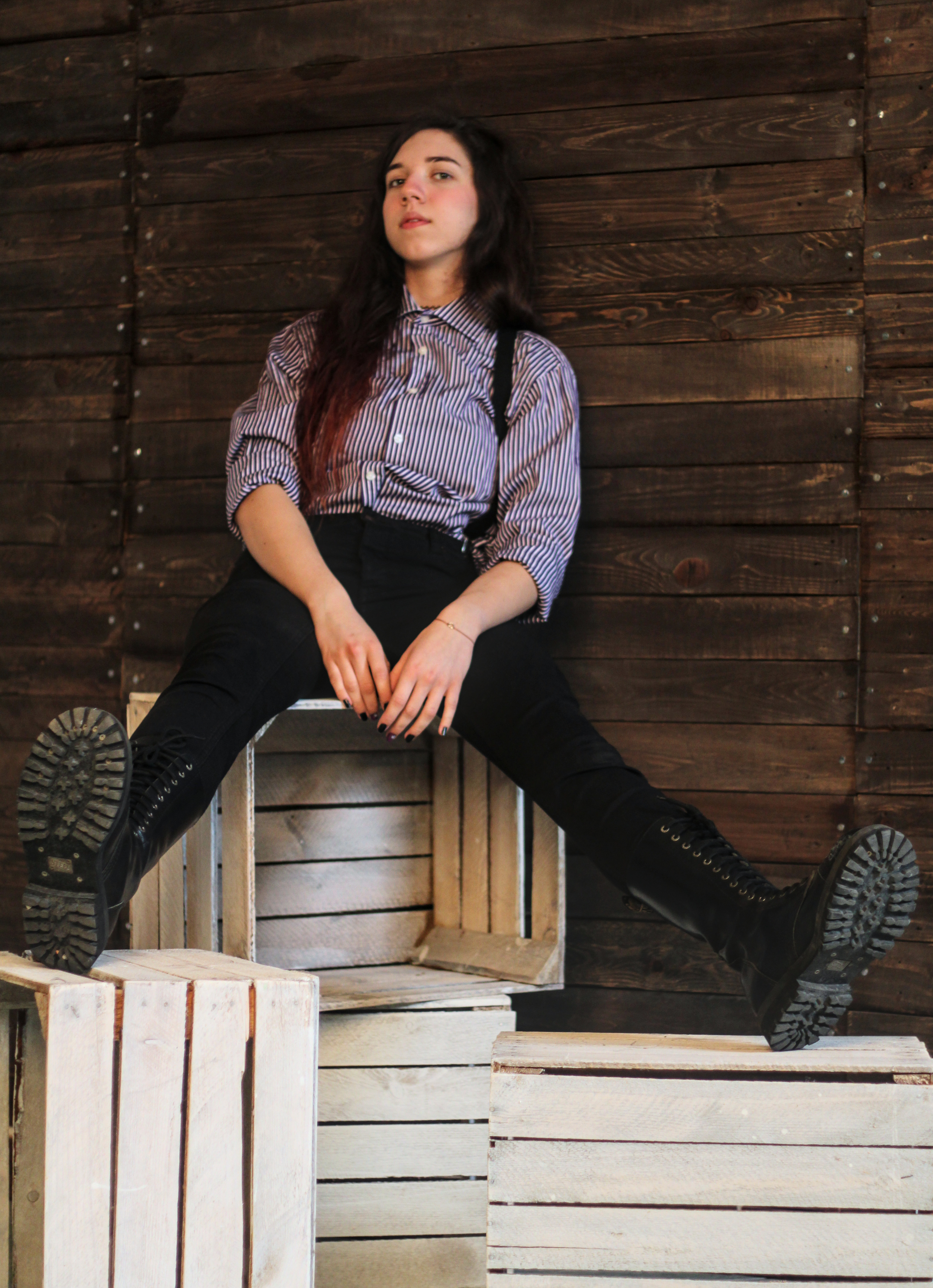 Portret młodej kobiety w paski koszuli i czarnych spodniach z szelkami, siedzącej na drewnianych skrzynkach na tle drewnianej ściany, z wyciągniętymi nogami w czarnych, sznurowanych butach.