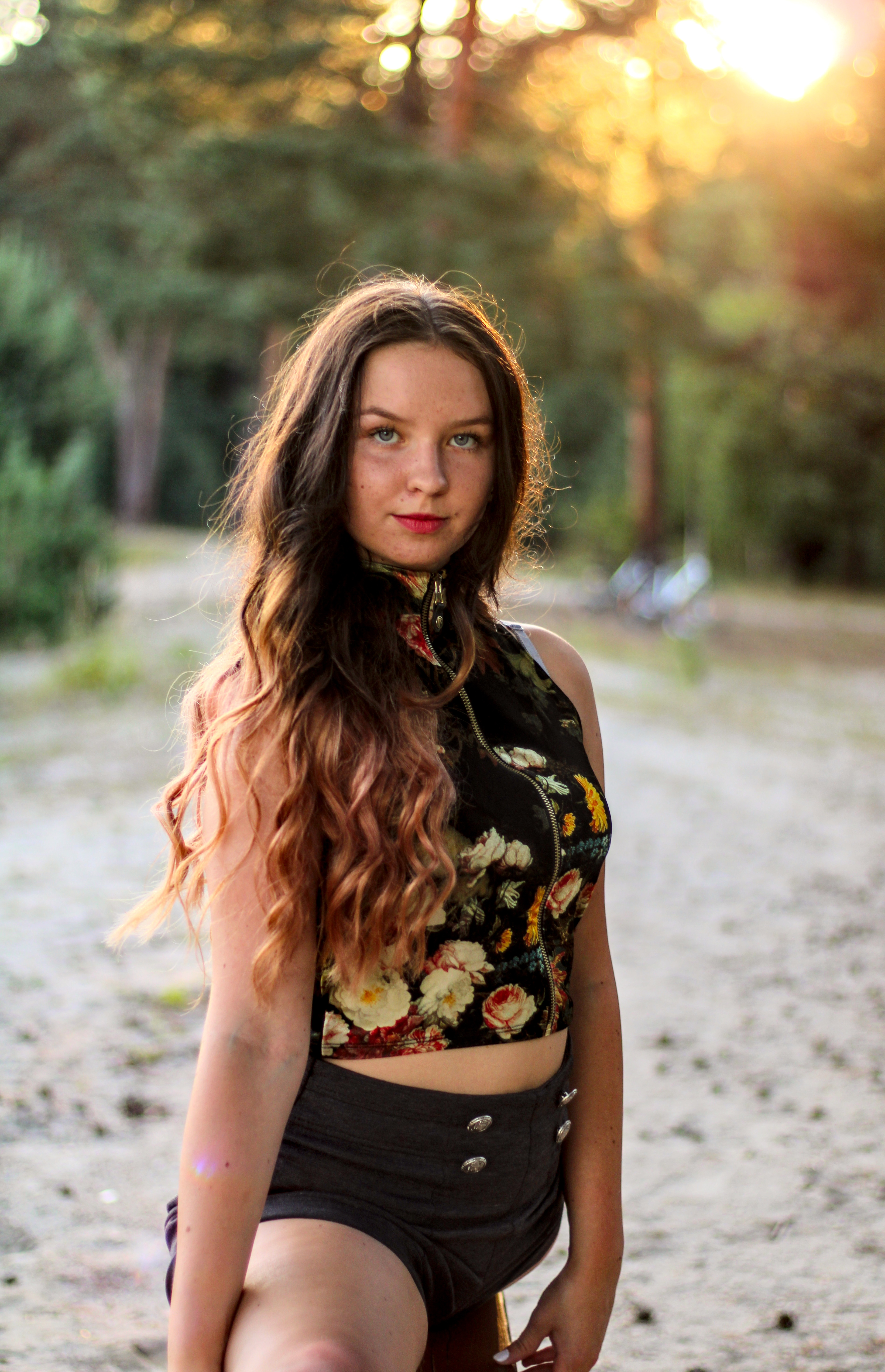Portret młodej kobiety z długimi, falowanymi włosami w kwiecistym topie i szortach na tle leśnym, oświetlonej popołudniowym słońcem.