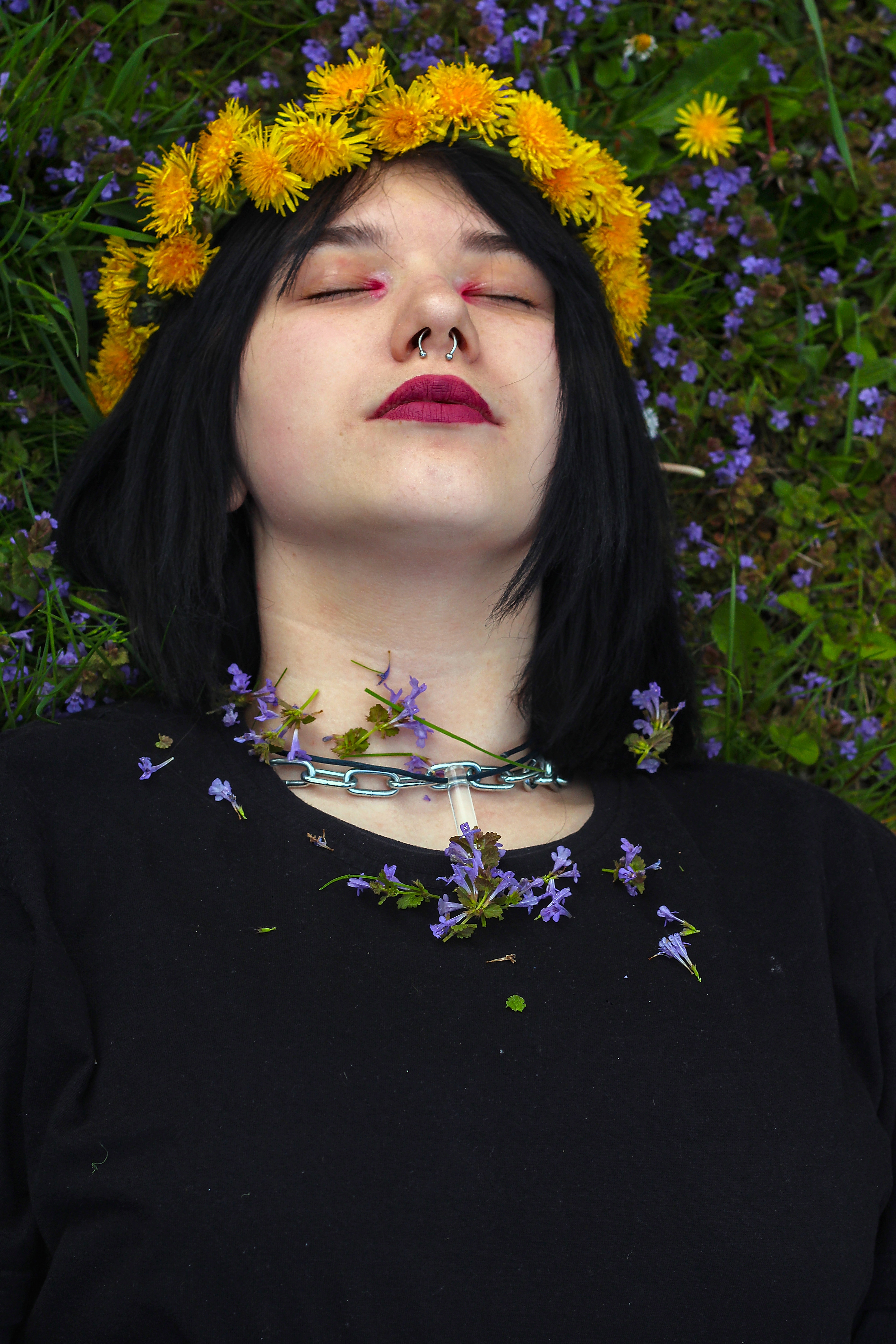 Młoda kobieta z kolczykiem w nosie i ciemną szminką, leżąca na trawie z wiankiem z mniszków lekarskich na głowie i łańcuchem z niebieskimi kwiatami na szyi.