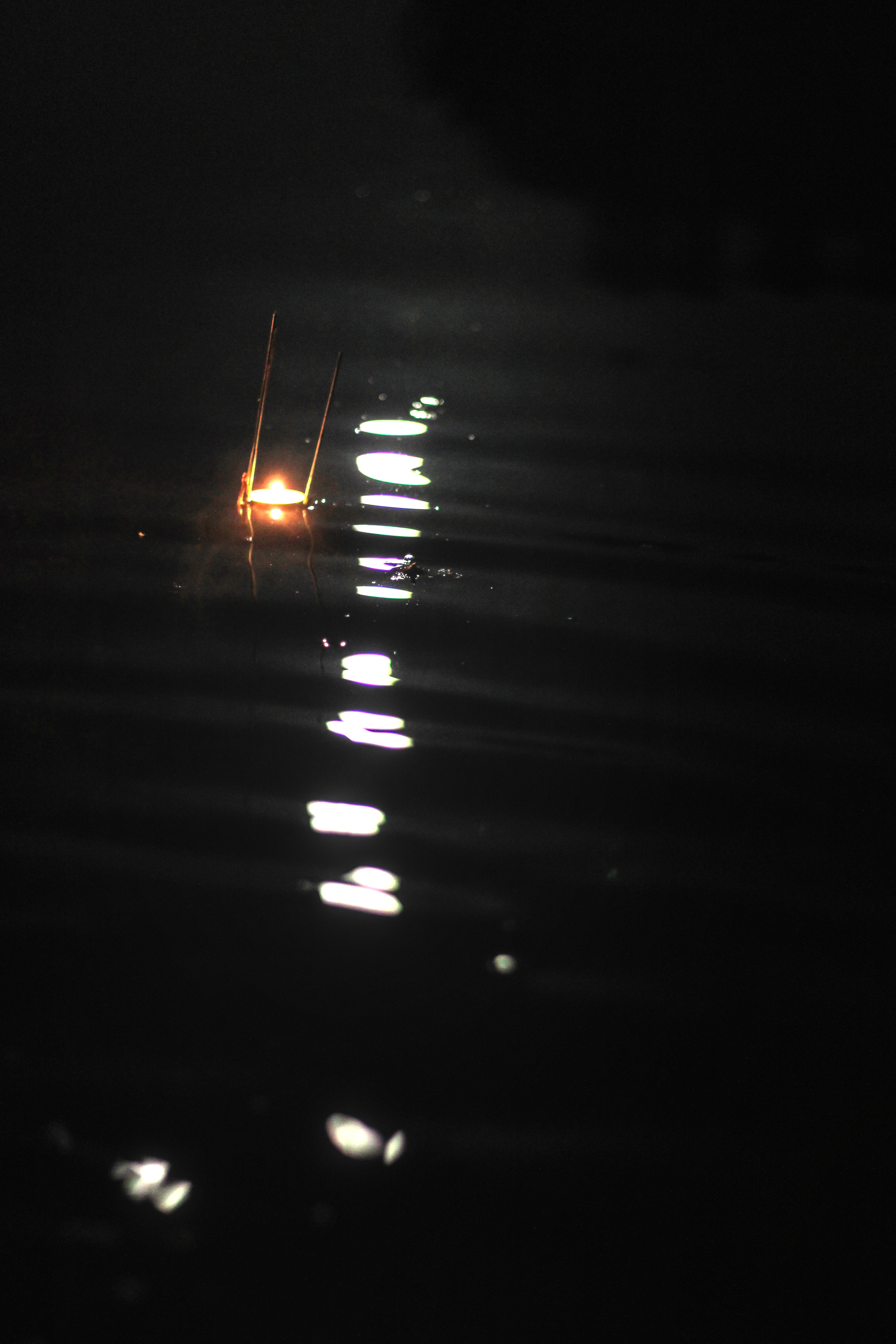 Płonąca świeca na wodzie w nocy, odbicie światła tworzy jasną ścieżkę, ciemne tło.