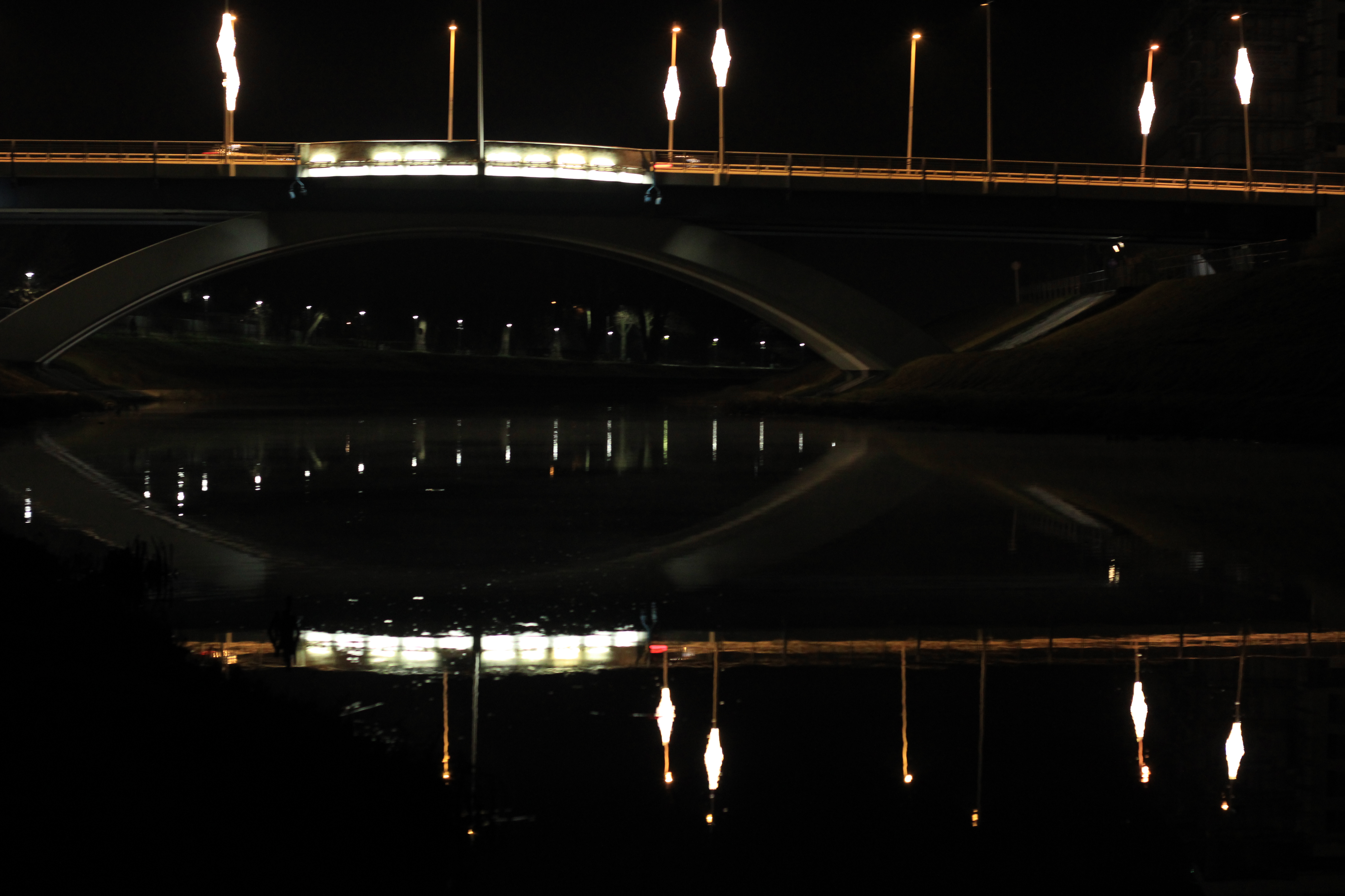 Nocny widok na most z oświetleniem odbijającym się w wodzie, z delikatnymi smugami świateł samochodów na górze.