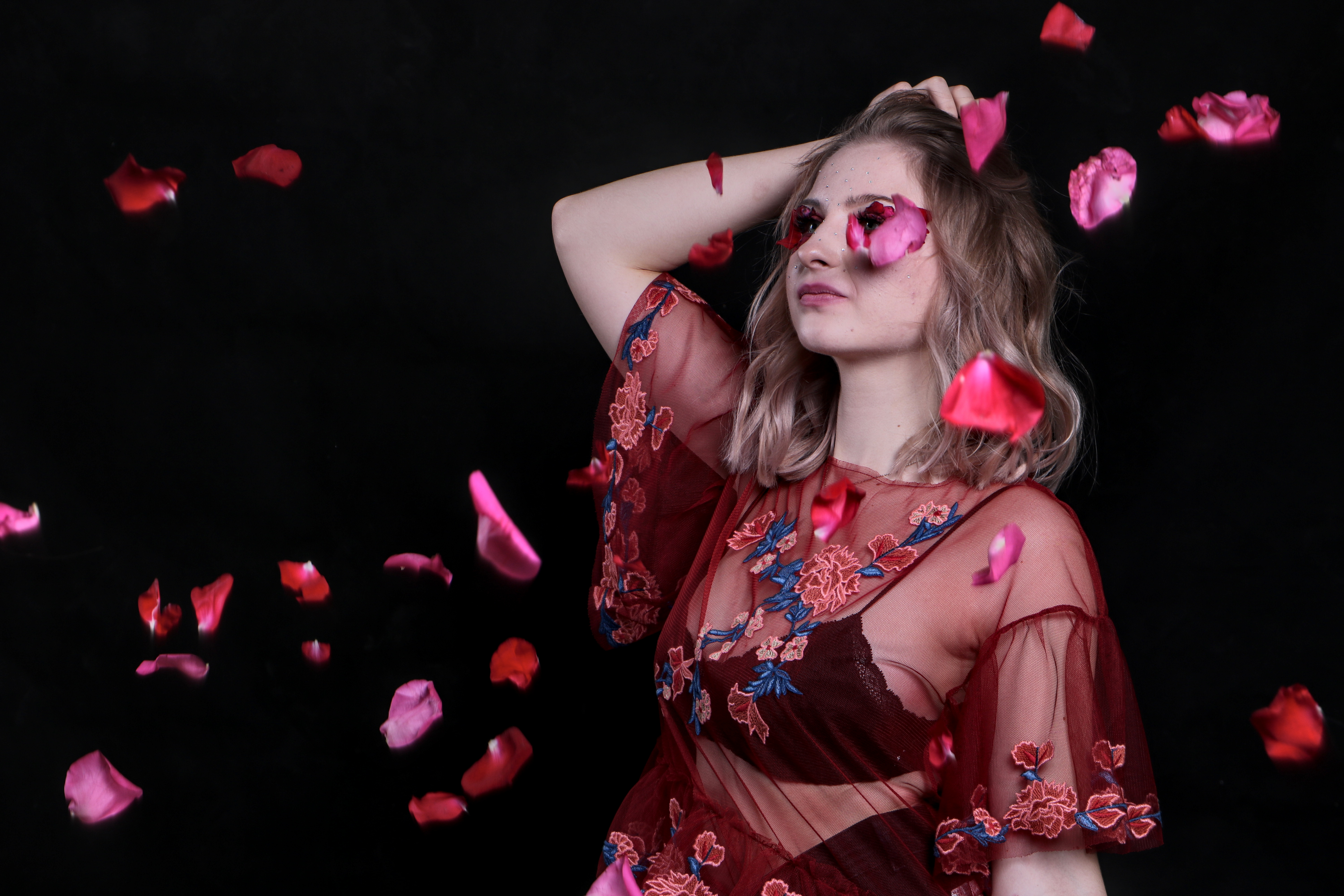 Młoda kobieta w przezroczystej, haftowanej bluzce pozuje z opadającymi płatkami róż na czarnym tle, delikatny makijaż i ręka we włosach.