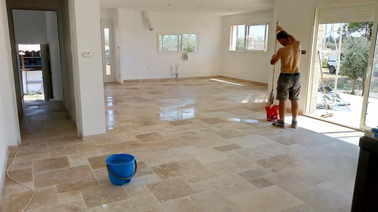 Mężczyzna myje nową, beżową podłogę z płytek o nieregularnym kształcie w przestronnym, jasnym wnętrzu domu. Widoczne wiadro z wodą i mop.