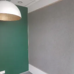 Pomieszczenie biurowe z zieloną ścianą i szarą ścianą pokrytą materiałem dźwiękochłonnym, oświetlone wiszącą lampą z białym kloszem i złotym wnętrzem.