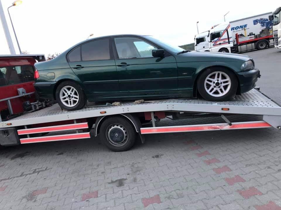 Zielony sedan BMW serii 3 na platformie lawety, widok z boku, w tle zaparkowane ciężarówki.