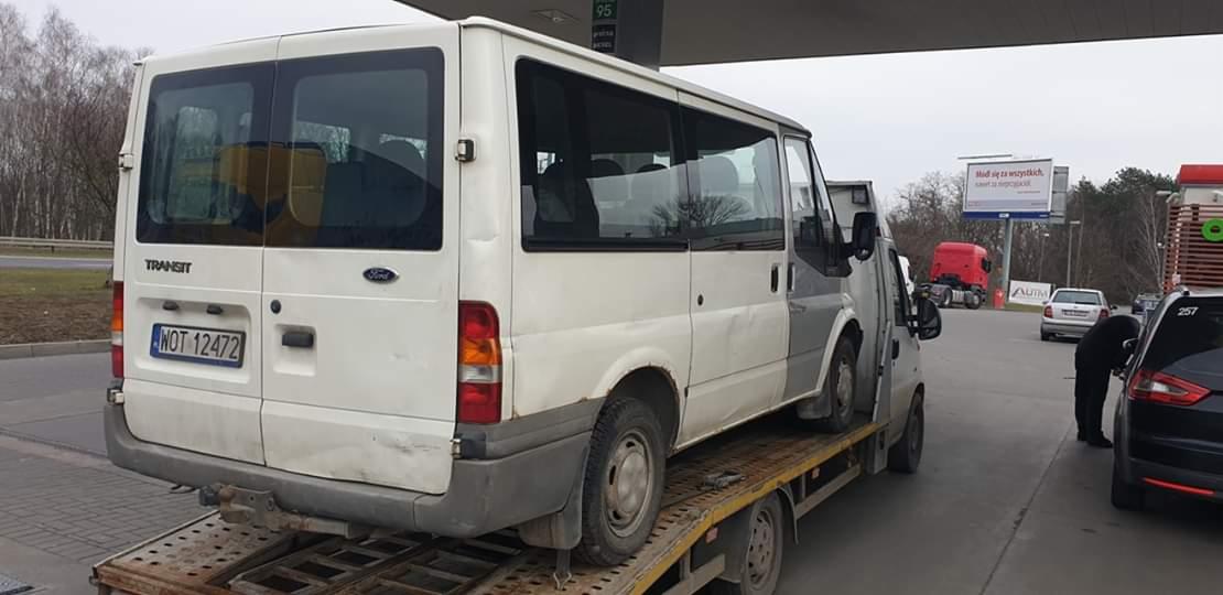 Uszkodzony biały Ford Transit z widocznymi śladami korozji transportowany na lawecie, na tle stacji benzynowej.