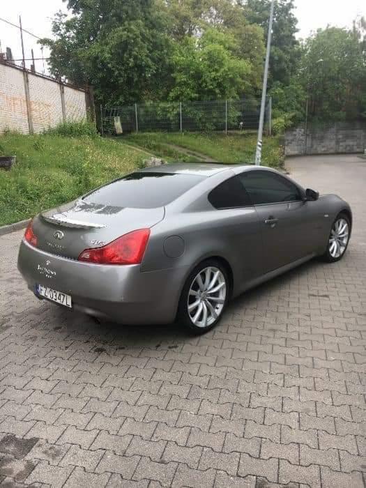 Szary samochód marki Infiniti G37 Coupe, zaparkowany na kostce brukowej, widok z tyłu i boku, z widocznymi chromowanymi felgami i przyciemnianymi szybami.