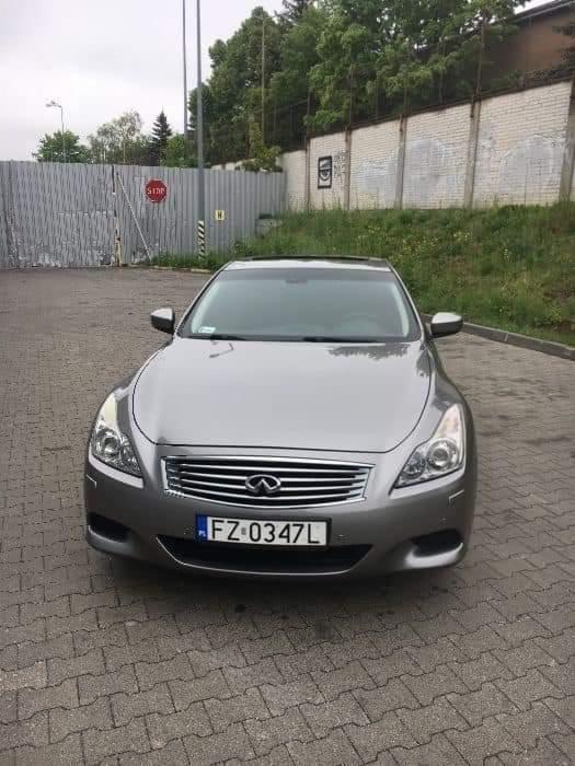 Szary samochód Infiniti G37 z polską tablicą rejestracyjną FZ 0347L, zaparkowany na brukowanym placu, na tle metalowego ogrodzenia i betonowego muru z graffiti.
