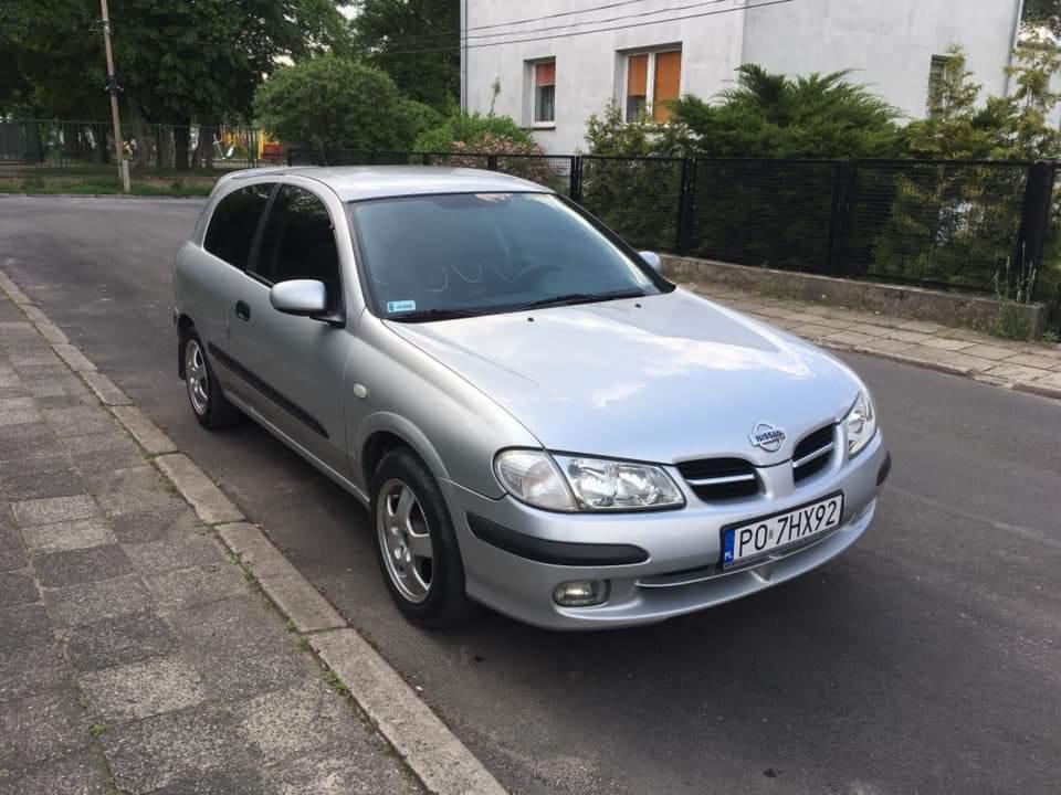 Srebrny Nissan Almera zaparkowany na ulicy, widoczne tablice rejestracyjne i uszkodzenia na szybie przedniej.