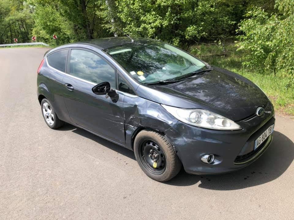 Uszkodzony, szary Ford Fiesta z przerysowanym bokiem i brakiem kołpaka, stojący na poboczu drogi.