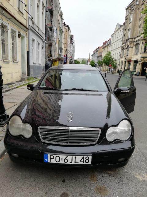 Czarny Mercedes z otwartymi drzwiami pasażera, zaparkowany na ulicy w centrum miasta z budynkami w tle.