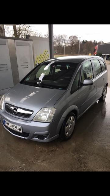 Srebrny Opel Meriva zaparkowany na mokrej powierzchni, widoczny numer rejestracyjny PM1-02UW, zarys drzew w tle.