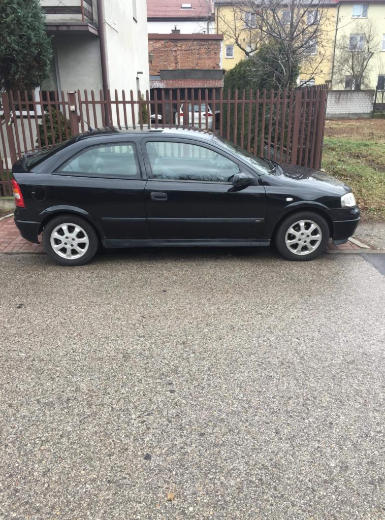 Czarny Opel Astra II Coupe zaparkowany na mokrym, szarym asfalcie przed brązowym ogrodzeniem i budynkami mieszkalnymi w tle.