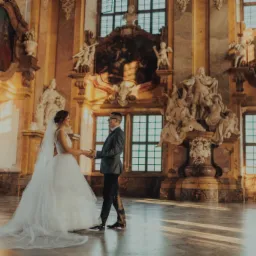 Elegancka para młoda trzymająca się za ręce w bogato zdobionym wnętrzu pałacowym, z marmurową posadzką i rzeźbami. Ciepłe, naturalne światło podkreśla uroczystość chwili.