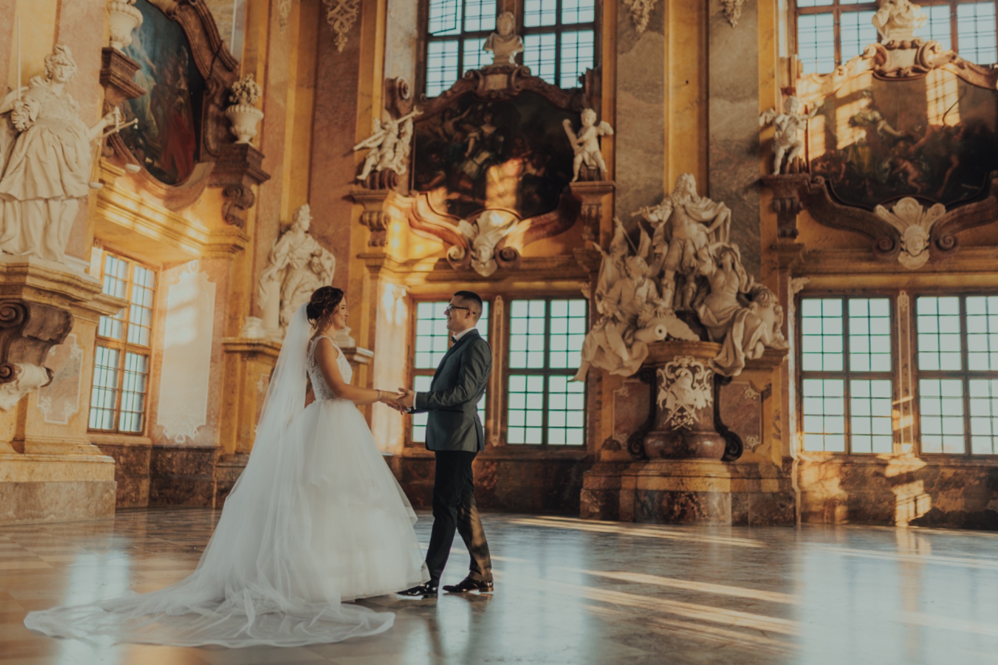Elegancka para młoda trzymająca się za ręce w bogato zdobionym wnętrzu pałacowym, z marmurową posadzką i rzeźbami. Ciepłe, naturalne światło podkreśla uroczystość chwili.