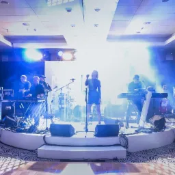 Zespół muzyczny na scenie podczas występu na evencie, wokalistka w krótkiej sukience, klawiszowcy po bokach, tło z nazwiskami artystów, oświetlenie sceniczne.