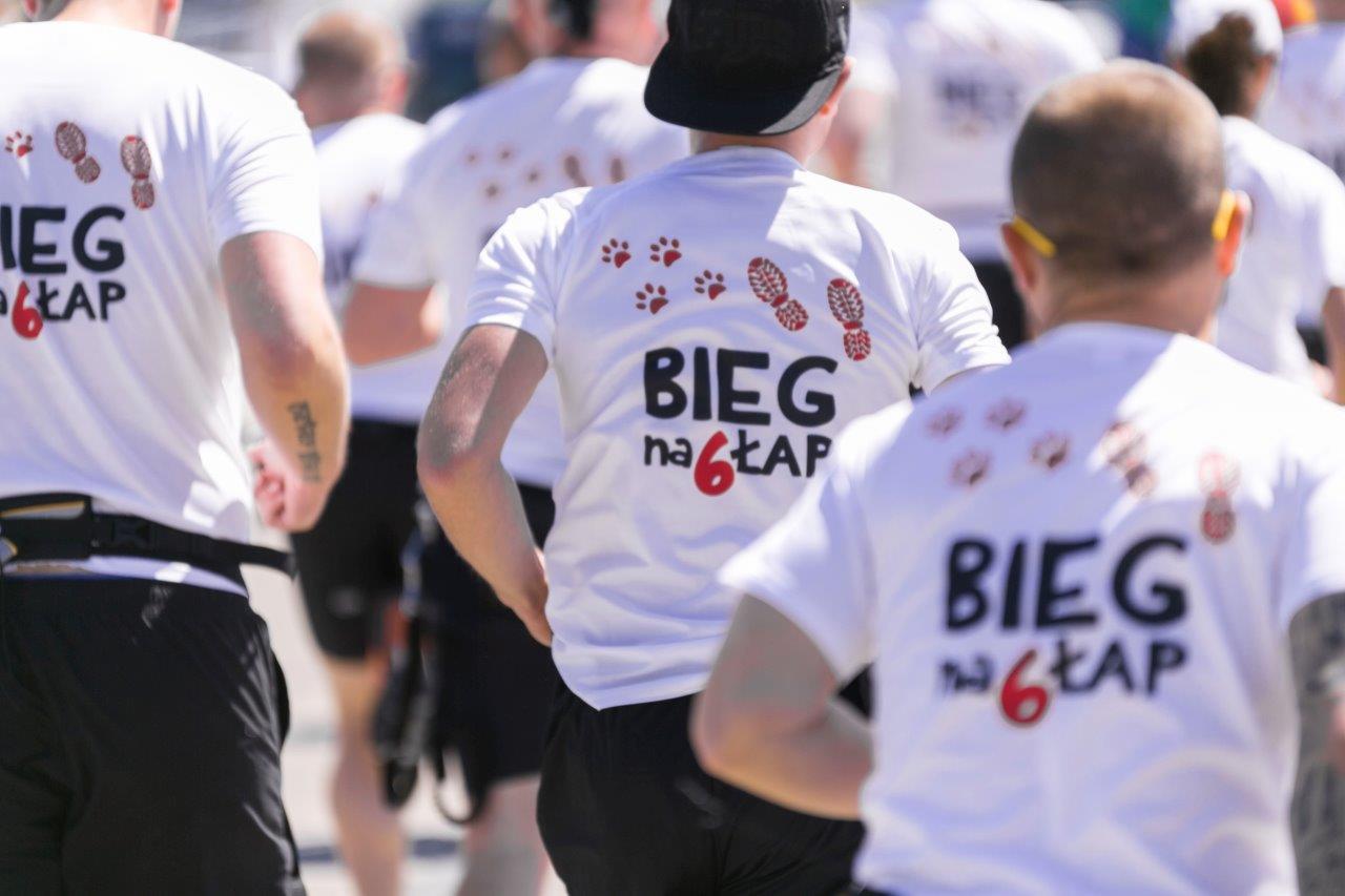 Uczestnicy biegu charytatywnego 'Bieg na 6 Łap' w białych koszulkach z nadrukiem, widok z tyłu, skupienie na koszulkach z motywem psich łap i odcisków butów.