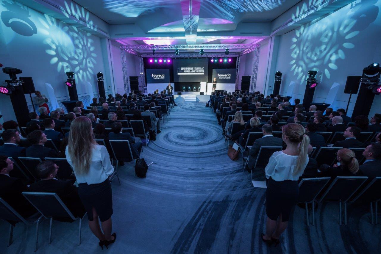 Sala konferencyjna wypełniona uczestnikami, scena z ekranem wyświetlającym nazwę firmy i prelegentów, oświetlenie sceniczne w kolorach fioletu i niebieskiego, widok z tyłu sali.