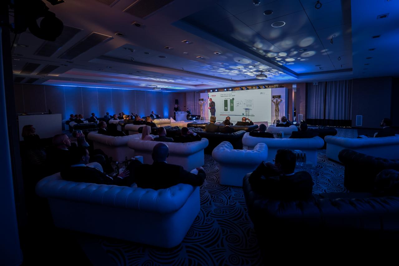 Konferencja w sali z niebieskim oświetleniem, uczestnicy siedzą na białych i czarnych sofach, prelegent prezentuje dane na ekranie.