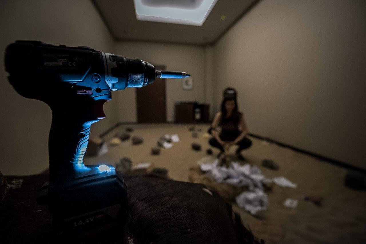 Akumulatorowa wiertarko-wkrętarka na pierwszym planie, w tle kobieta w kucki sprzątająca podłogę z resztek materiałów budowlanych w pustym pomieszczeniu z oświetleniem sufitowym.