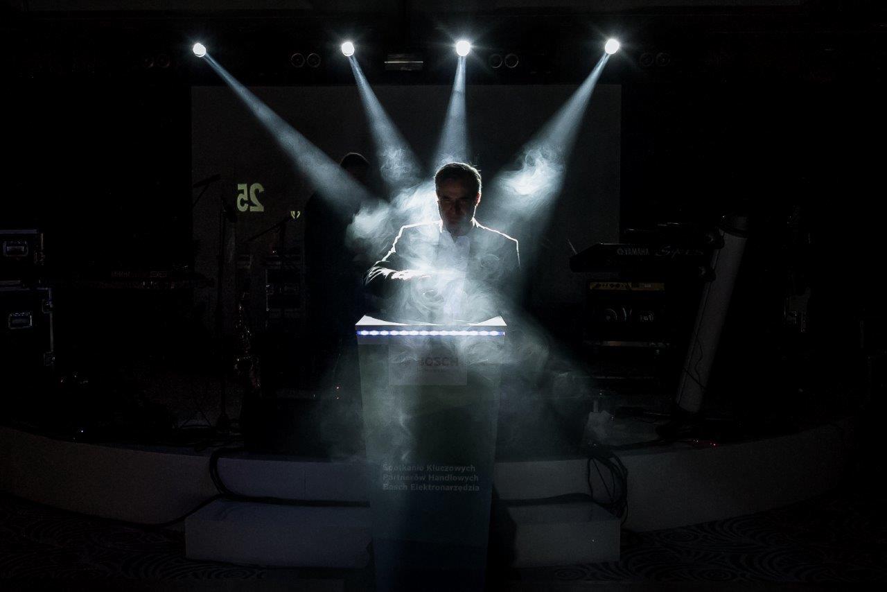 Mężczyzna w garniturze za podświetlonym pulpitem na scenie w oparach dymu i reflektorów.