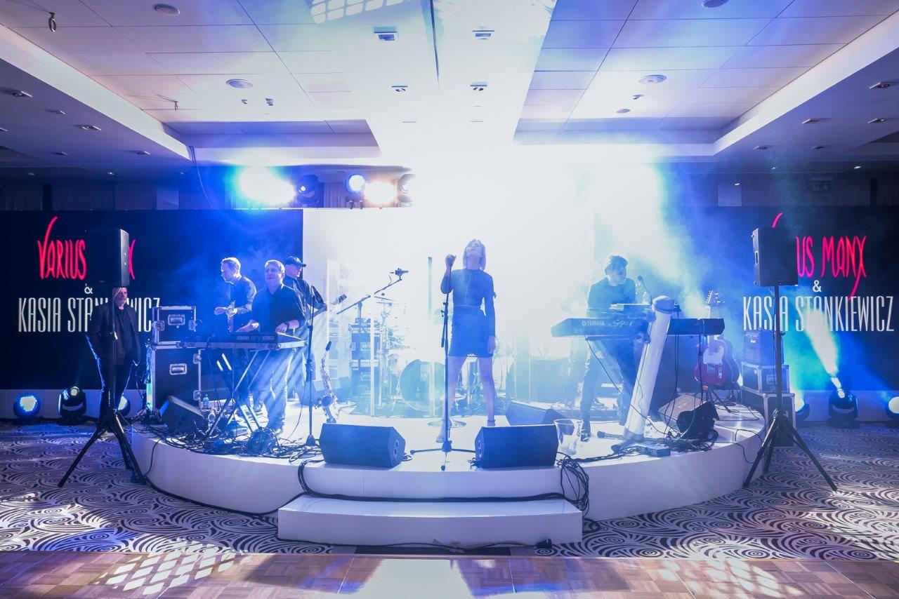 Zespół muzyczny na scenie podczas występu na evencie, wokalistka w krótkiej sukience, klawiszowcy po bokach, tło z nazwiskami artystów, oświetlenie sceniczne.