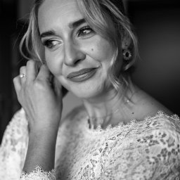 DOMINIKA UŁAN KAWALERIA FOTOGRAFII - Czarno-białe zbliżenie panny młodej w koronkowej sukni, poprawiającej kolczyk, z pierścionkiem zaręczynowym na palcu, spoglądającej w lewo.