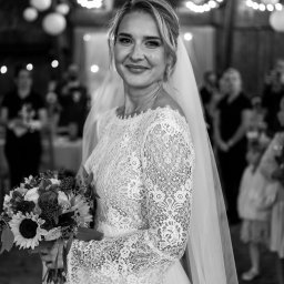 DOMINIKA UŁAN KAWALERIA FOTOGRAFII - Portret uśmiechniętej panny młodej w koronkowej sukni z bukietem słoneczników, welonem i delikatną biżuterią na tle sali z dekoracyjnym oświetleniem.