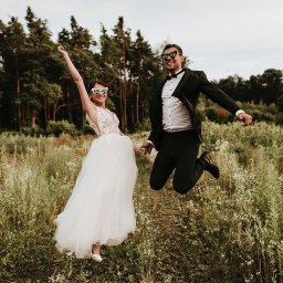 DOMINIKA UŁAN KAWALERIA FOTOGRAFII - Uśmiechnięta para młoda w skoku na tle łąki i lasu, oboje w okularach w kształcie serc z napisem 'bride' i 'groom'.