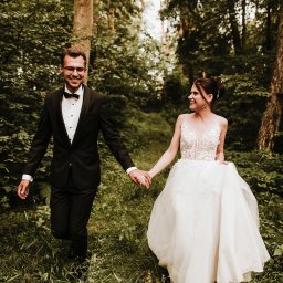 DOMINIKA UŁAN KAWALERIA FOTOGRAFII - Uśmiechnięta para młoda, on w czarnym smokingu i okularach, ona w białej sukni z koronkową górą, trzymają się za ręce idąc przez zarośnięty las.