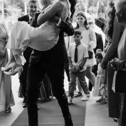 Pan młody niesie uśmiechniętą pannę młodą na rękach po czerwonym dywanie, w tle goście weselni i dziecko w krawacie.