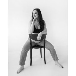 DOMINIKA UŁAN KAWALERIA FOTOGRAFII - Czarno-białe zdjęcie kobiety w ciąży siedzącej tyłem na krześle, w dżinsach i topie, z ręką podpartą pod brodą, zamyślony wzrok skierowany w lewo.