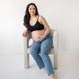 DOMINIKA UŁAN KAWALERIA FOTOGRAFII - Kobieta w ciąży, w czarnym topie i jeansach, siedzi na krześle na białym tle, trzymając ręce na brzuchu i uśmiechając się.