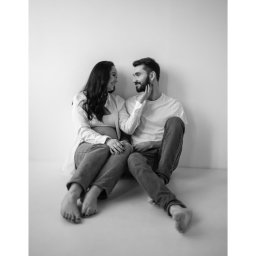DOMINIKA UŁAN KAWALERIA FOTOGRAFII - Czarno-białe zdjęcie pary siedzącej na podłodze: kobieta w ciąży w rozpiętej białej koszuli i jeansach, mężczyzna z brodą w białym t-shircie i jeansach, delikatnie dotyka jej twarzy, oboje patrzą...