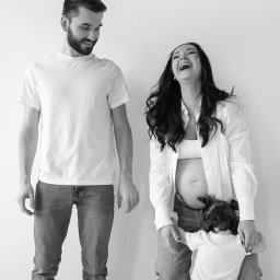 DOMINIKA UŁAN KAWALERIA FOTOGRAFII - Czarno-białe zdjęcie szczęśliwej rodziny w Niepołomicach: uśmiechnięty ojciec, śmiejąca się matka w ciąży i mała dziewczynka przytulająca się do jej brzucha.