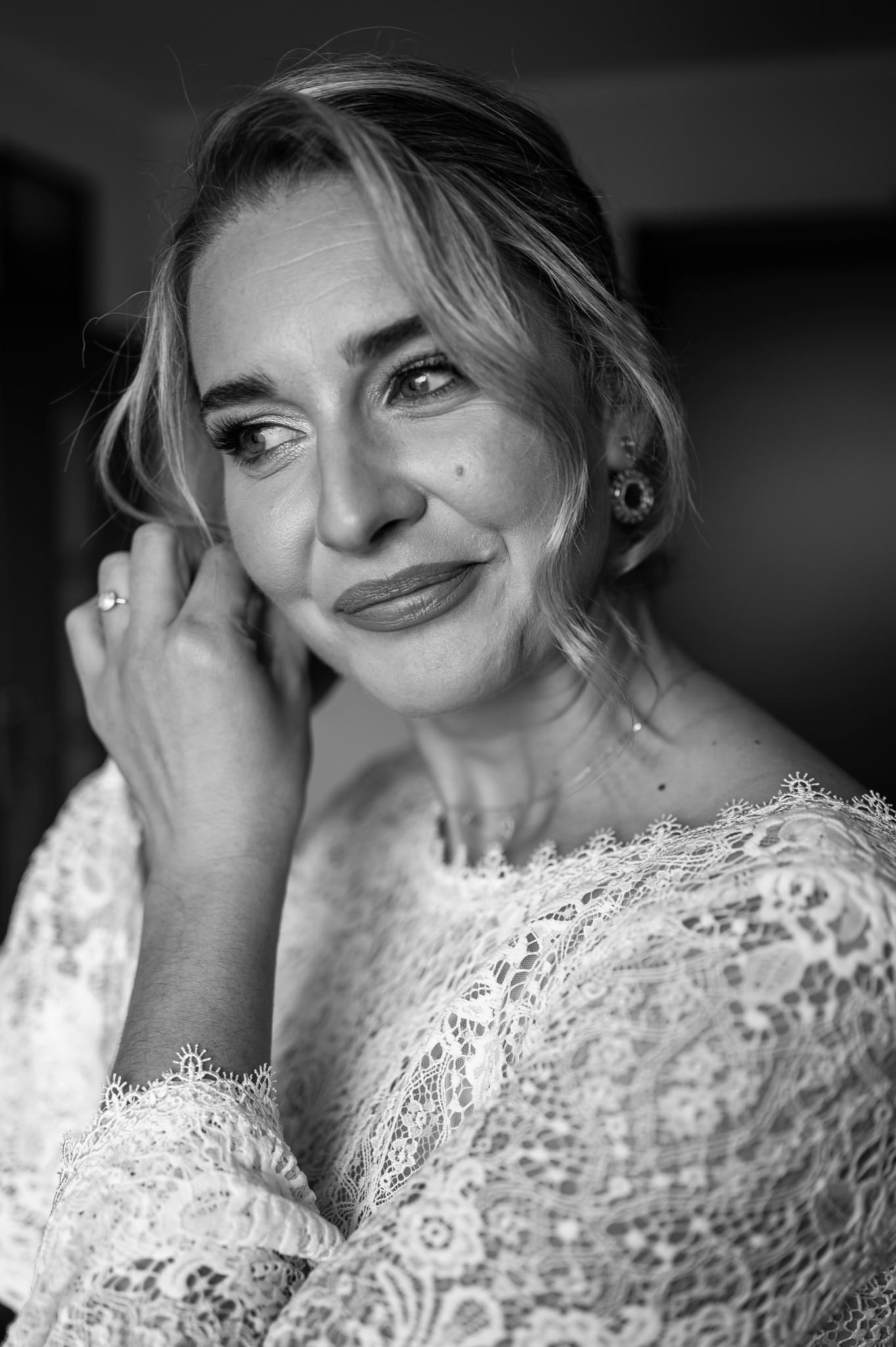 Czarno-białe zbliżenie panny młodej w koronkowej sukni, poprawiającej kolczyk, z pierścionkiem zaręczynowym na palcu, spoglądającej w lewo.